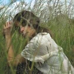 Claire is crouching eyes closed in the middle of very tall grasses which are taller than her in this position. She looks peaceful and immersed, totally in tune with natural world that she is part of.